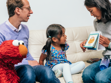 A small child sits with Elmo as their family reads flashcards.