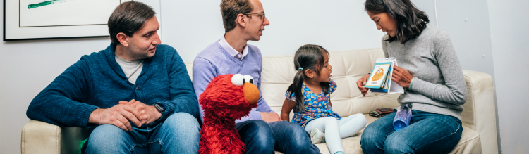 A small child sits with Elmo as their family reads flashcards.