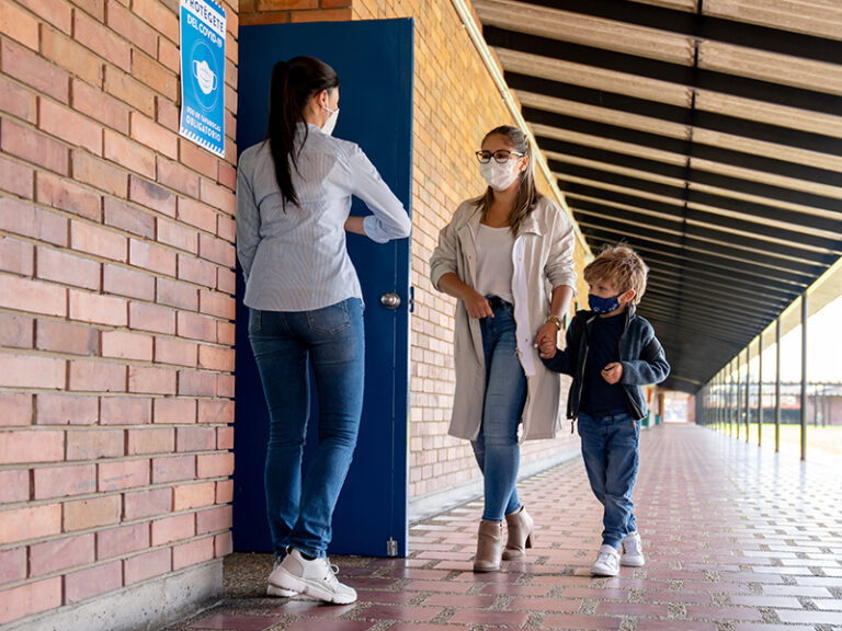 An adult holds the hand of a child as the walk through a door held open by another adult wearing a mask.