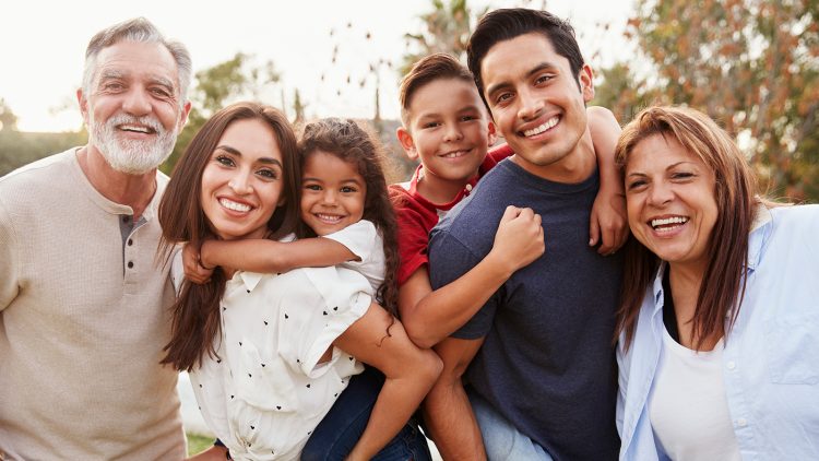 Six family members standing outside, including two children getting piggyback rides with adults, all are smiling.