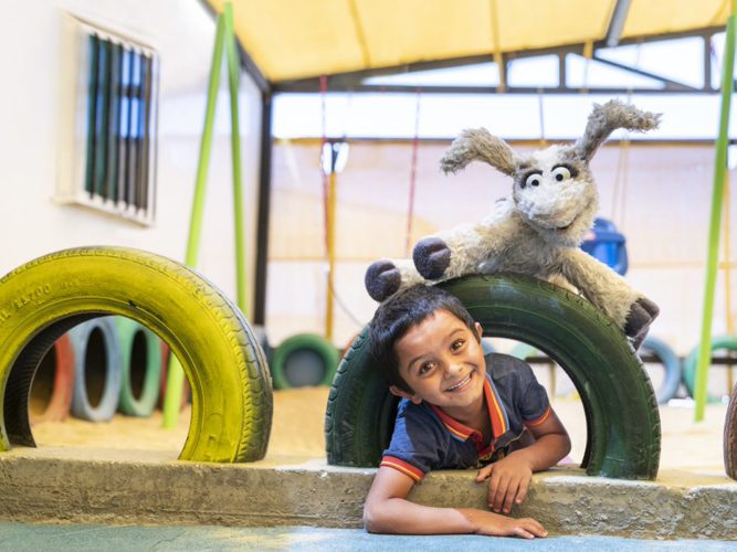 Ma'Zooza (a goat muppet) playing with a child on a playground.