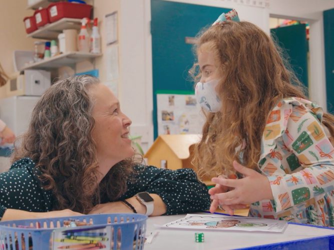 A teacher kneels down to speak to a student at their desk