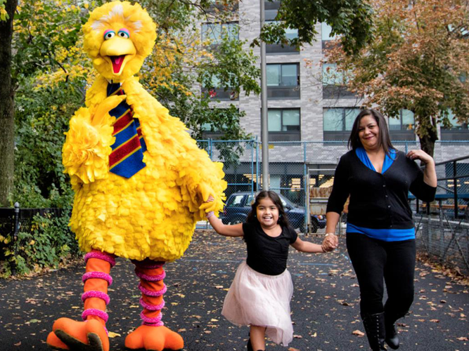 Big Bird walks towards us, holding the hand of a young child who's holding the hand of an adult.