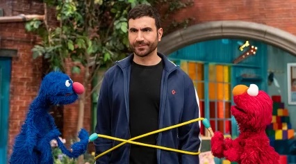 Elmo and Grover wrap ropes around actor Brett Goldstein.