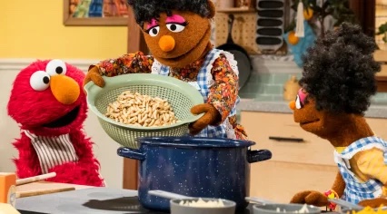 Grandma Nell holds up a colander next to a pot. Elmo and Gabrielle.