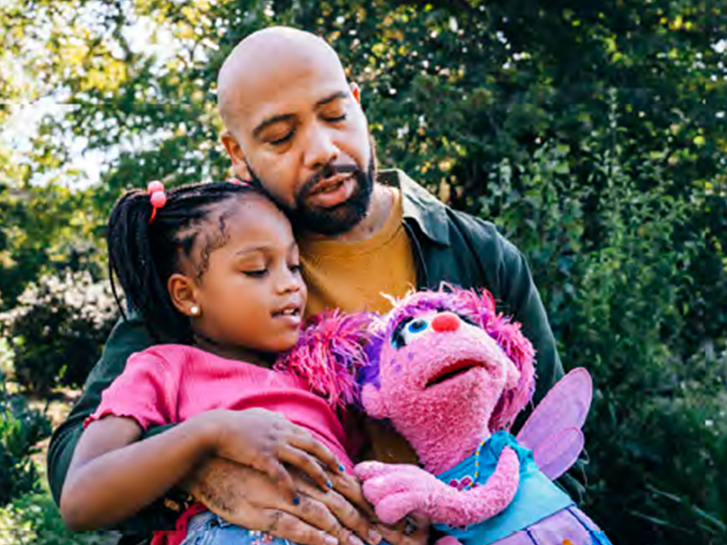 A caregiver and a child are hugging Abby Cadabby.