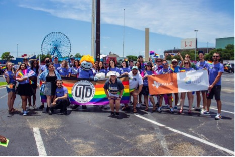 A group of students and staff stand with a UT Dallas progress rainbow flag and a QUTD sign.