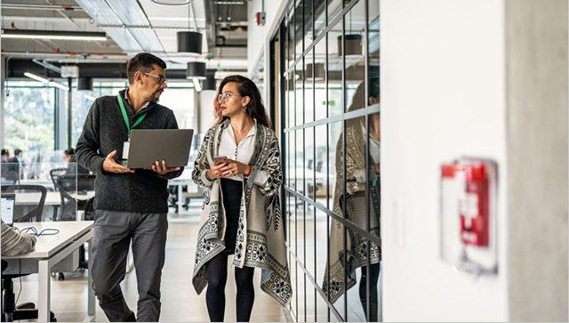 Two people in conversation walk through an office hallway--one person holds a laptop