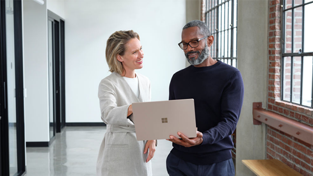 Photo of two people standing in a hallway and in conversation while looking at a laptop.