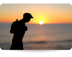 Closeup of a paper cutout in the shape of a young man, wearing a flat cap and carrying a backpack, in front of the ocean