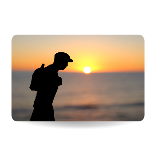 Closeup of a paper cutout in the shape of a young man, wearing a flat cap and carrying a backpack, in front of the ocean