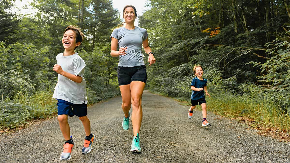 Active Runner Mom Running On Trail With Two Kids On Either Side Of Her, Smiling And Enjoying The Outdoors