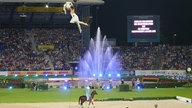 Die Eröffnungsfeier im Reitstadion: Über einer riesigen Wasserfontäne schwebt eine Akrobatin an einem Seil. Die Besuchertribünen sind voll besetzt.