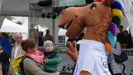 Das Maskottchen der Reit-EM begrüßt ein kleines Kind, das sich in den Armen seiner Mutter befindet.