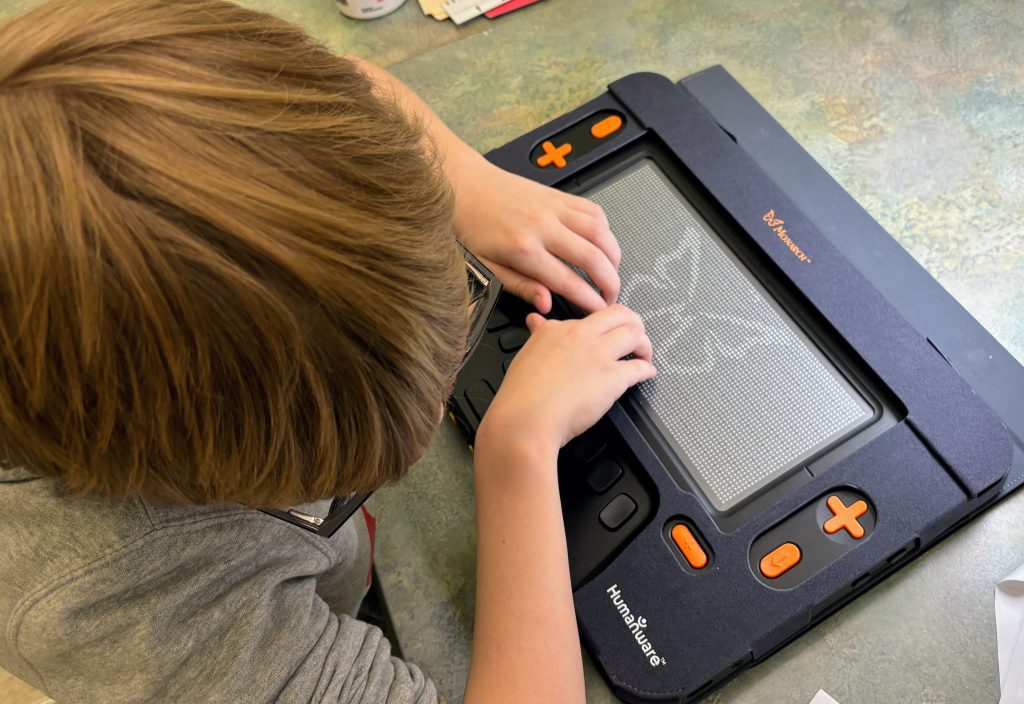 Overhead shot of blonde boy in glasses exploring a tactile graphic of a Monarch butterfly rendered on the Monarch's surface.