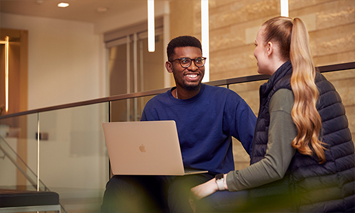 Chidi seated with a MacBook on his knees, speaking with a colleague.