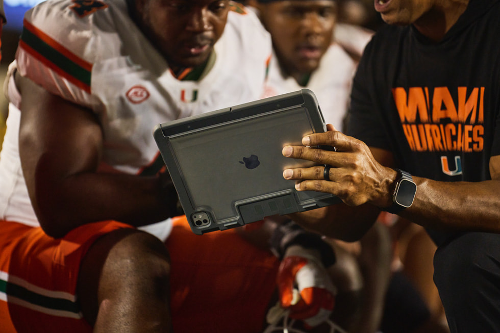 A Miami coach reviews footage on iPad with players on the sideline during the game.