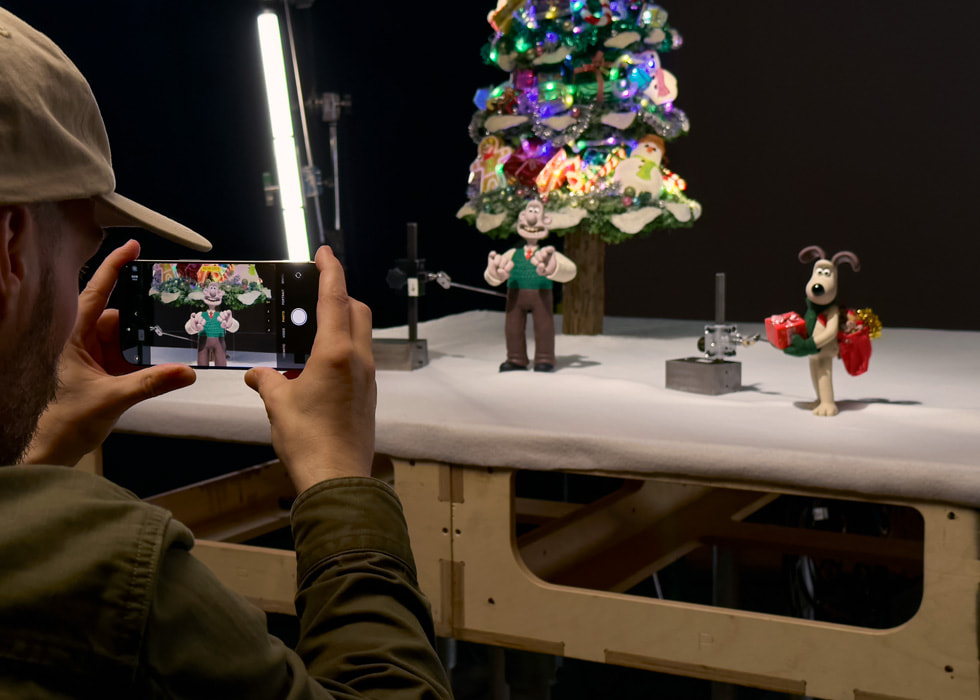 As part of the stop-motion animation process, a person holding iPhone 16 Pro Max captures stills of miniature clay Wallace & Gromit figures in front of a Christmas tree.