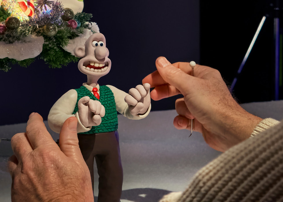 As part of the stop-motion animation process, a person holding iPhone 16 Pro Max captures stills of miniature clay Wallace & Gromit figures in front of a Christmas tree.