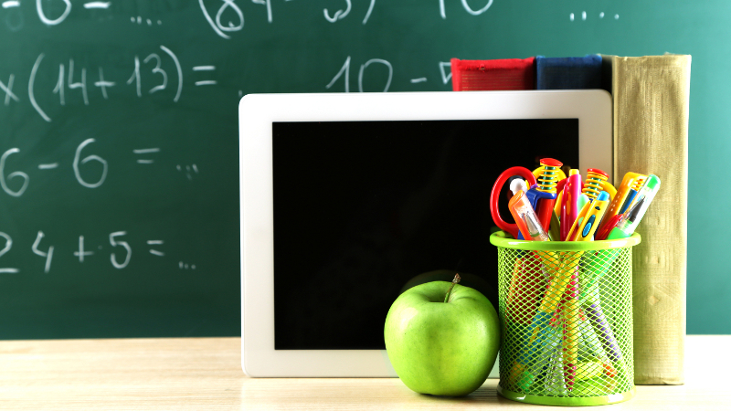 Bücher, Tablet, Stifte und Apfel auf einer Tischplatte vor einer mit Rechenaufgaben beschriebenen Schultafel.