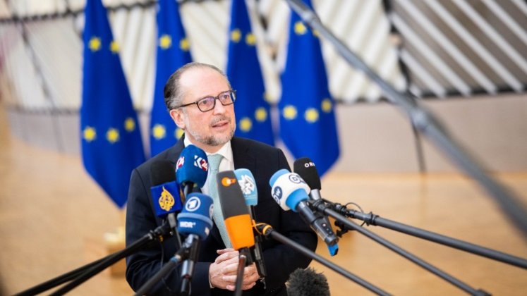 Außenminister Alexander Schallenberg steht in einem hellen Raum vor mehreren blau-gelben EU-Flaggen. Schallenberg spricht in Mikrofone mehrerer internationaler Medien, die vor ihm aufgestellt sind, während er freundlich und konzentriert wirkt. Die Szene vermittelt eine Pressekonferenz oder ein Statement in einem diplomatischen Umfeld.