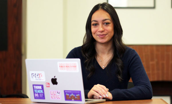 MacKenzie Hilton sits at a desk behind a laptop covered in BUSPH stickers, she has long dark hair and is wearing a navy blue long sleeve