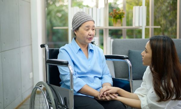 Cancer patient woman wearing head scarf sitting on wheelchair talking to her supportive daughter indoors, health and insurance concept.