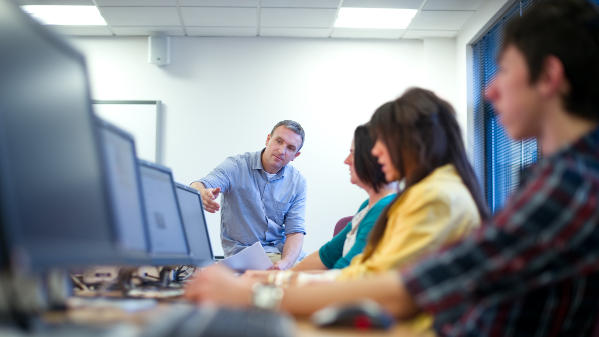 A group of students sitting at computer workstations while an instructor points at a screen, guiding them in a collaborative learning session.