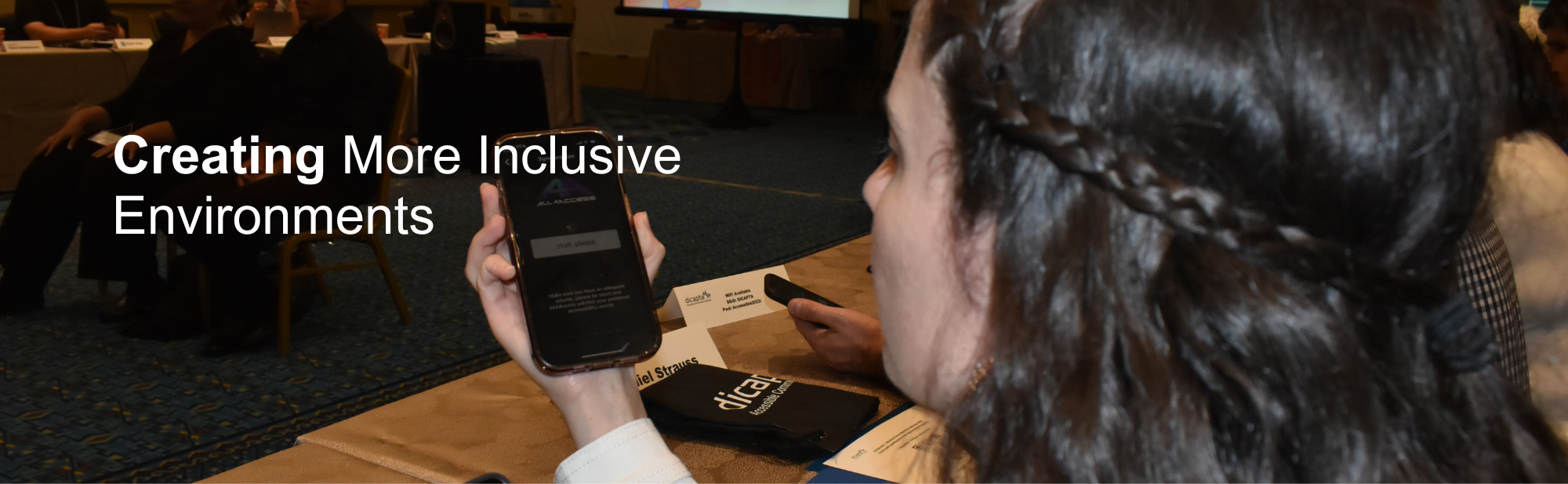 Text: Creating more inclusive environments. Background image: Conference room. A woman holds a phone that shows the All4Access app.