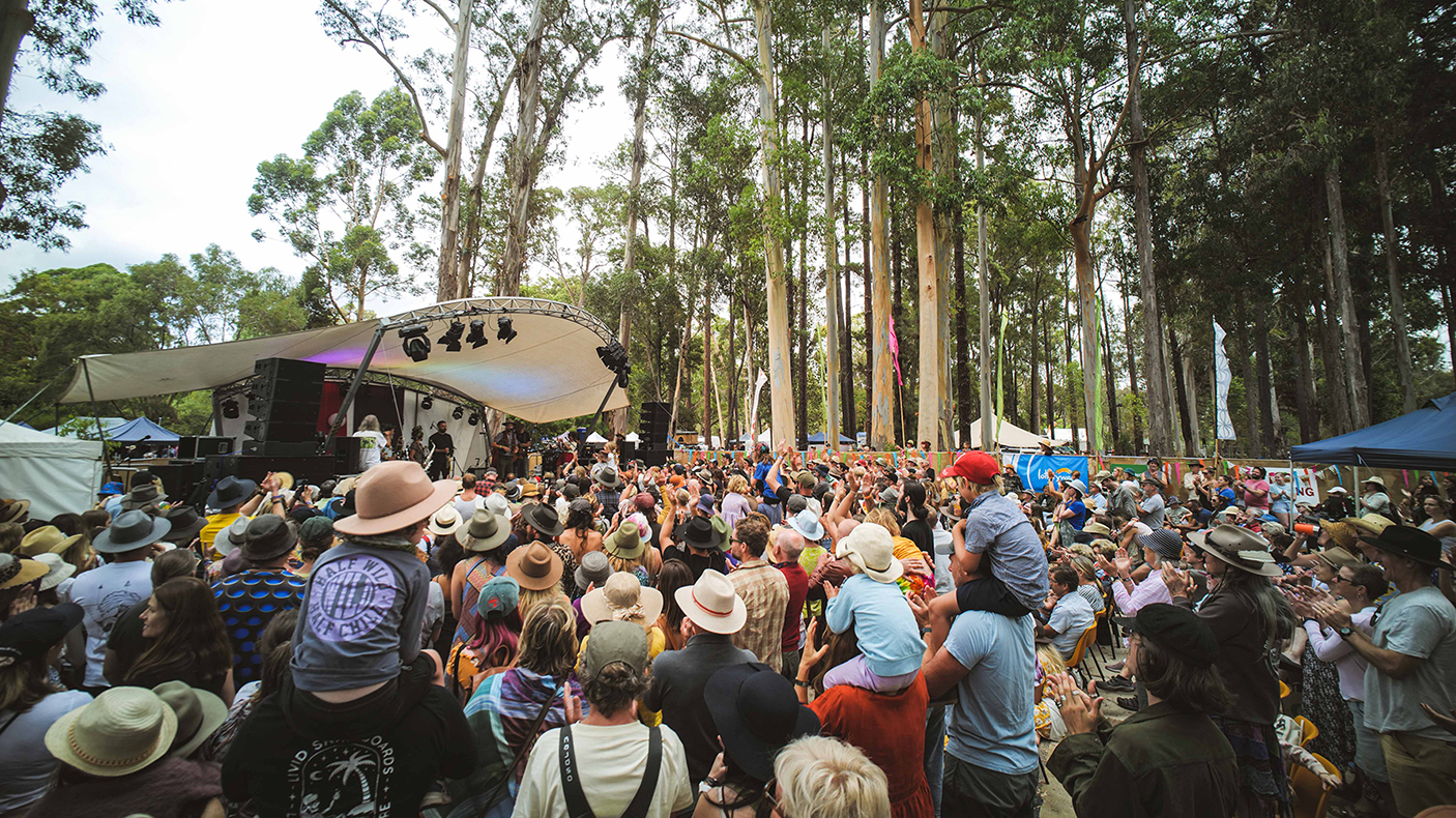 A large group of people watch the stage at a music festival in the forest.