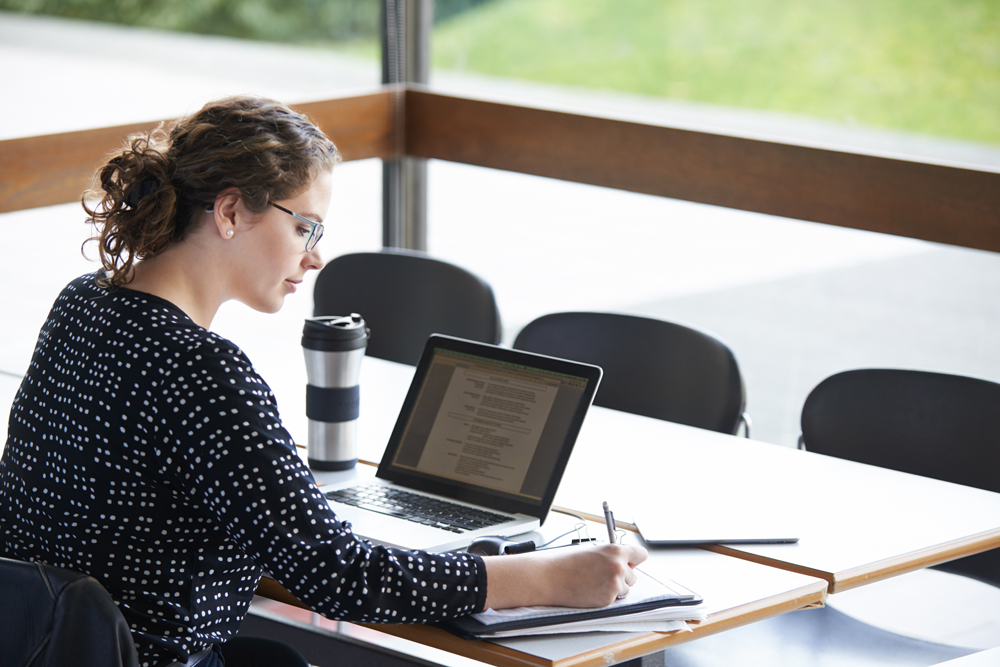 A woman woirking at a desk with a laptop and writing pad.