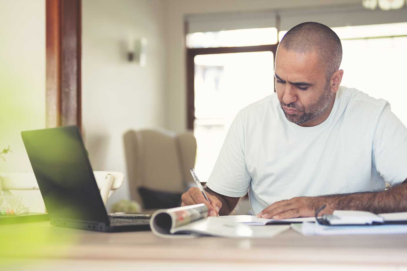 Mature man working at home. He has a laptop and is writing on a document at the table.