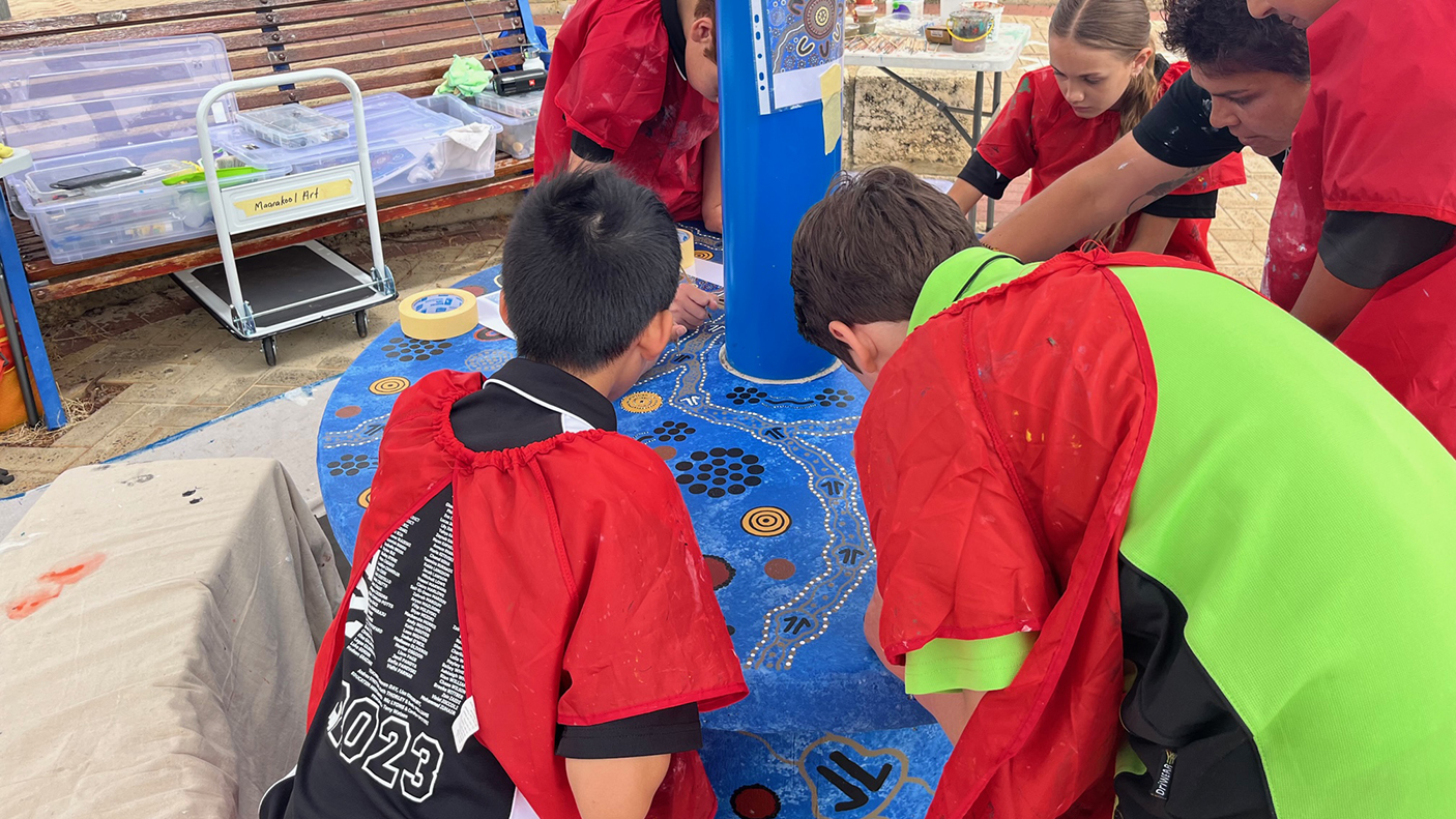 A group of school children painting Aboriginal designs on a circular artwork, together with an Aboriginal artist.