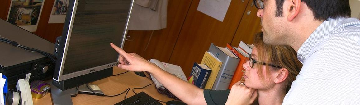 Woman sitting at a desk pointing to a text on a computer screen and a man looking over her shoulder