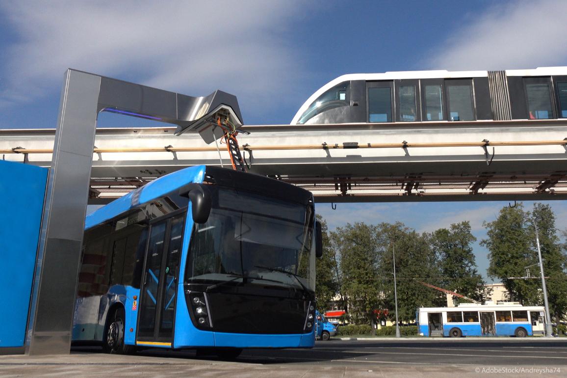 Ein blauer Elektrobus an einer Ladestation, während im Hintergrund eine moderne Einschienenbahn vorbeifährt.