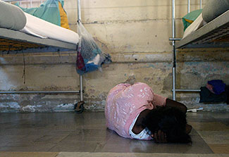 Person lies on floor curled into ball facing away from camera next to metal bed frames in an institution