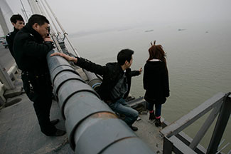 Woman facing away from camera stands on edge of tall bridge above water, rescue workers in background, her boyfriend reaches out to her