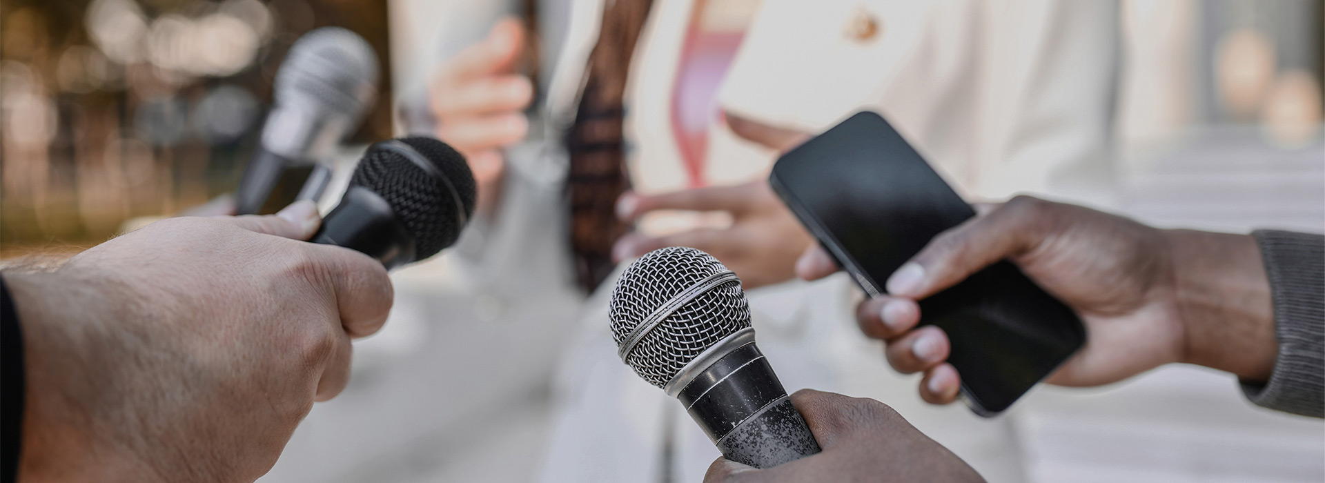 Hands holding out various microphones to a gesturing person in the background.