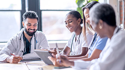 Group of four young doctors sitting happily at conference table discussing successful treatment with digital patient file on tablet