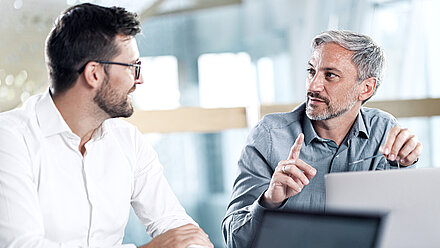 Front shot of two men sitting at a conference table in a bright, modern office, talking animatedly in front of a laptop: The man on the left is in his thirties, brunette and wears a beard, glasses and a white shirt and looks enthusiastically at his colleague (on his right); the man on the right is middle-aged, wears gray hair, a gray, short beard and a dove-blue shirt with small white dots and is explaining something important to his colleague, pointing upwards with his right index finger  Translated with DeepL.com (free version)