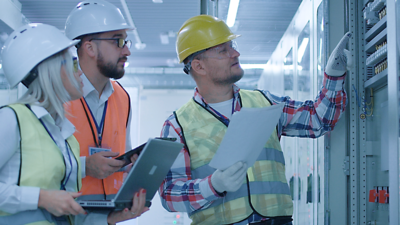 Three employees of a utility company wearing high-visibility vests and helmets are standing at a switchboard and power fuses; on the far right is a middle-aged blond man with a yellow helmet, protective gloves and a circuit diagram in his hand, showing the other two which settings they need to make; to their left is a younger brunette employee with glasses, a beard and a white helmet with a clipboard and a young blond female employee with a white shirt, high-visibility vest and laptop in her hand, listening attentively