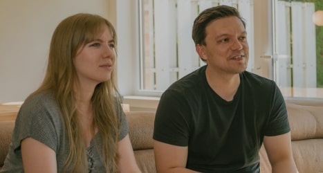 Pareja sentada en un sofá jugando videojuegos juntos en una sala luminosa con grandes ventanas.