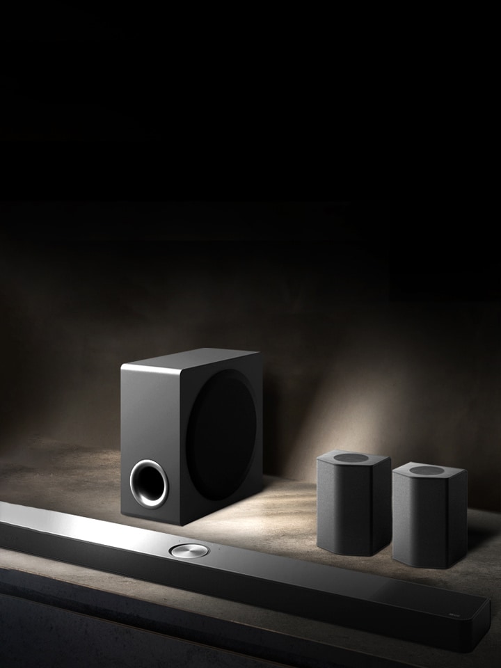 LG Soundbar, rear speakers, and subwoofer are placed within an angled perspective on a brown wooden shelf in a black room, enveloped in darkness with light only casting over the sound system.