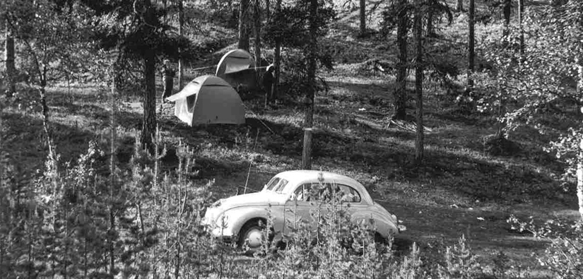 Two tents in the middle of the forest, an old car parked in the forest in the foreground.