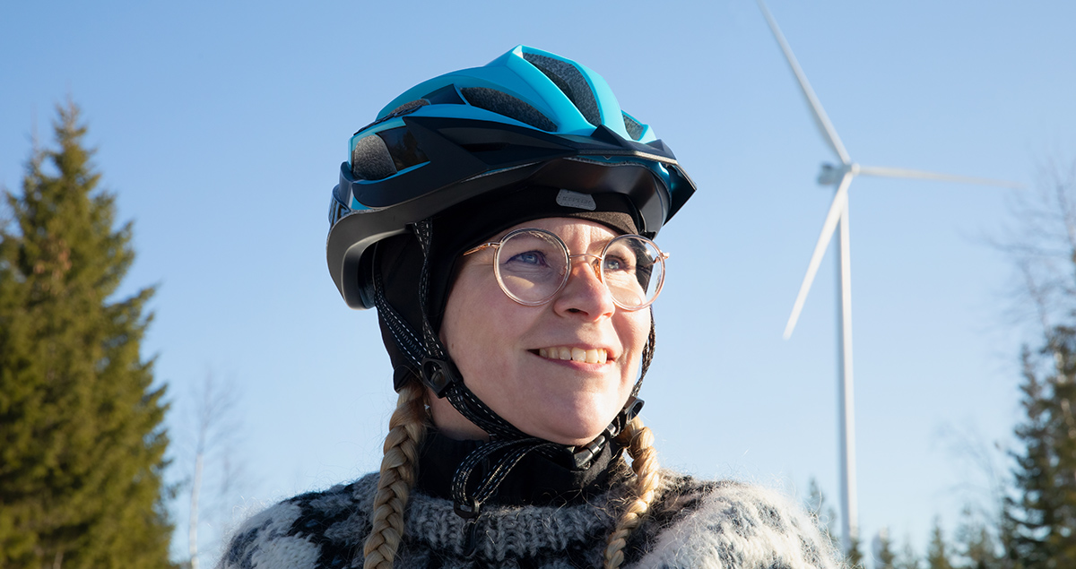 A woman in a close-up with a bicycle helmet and a wind power mast can be seen behind.