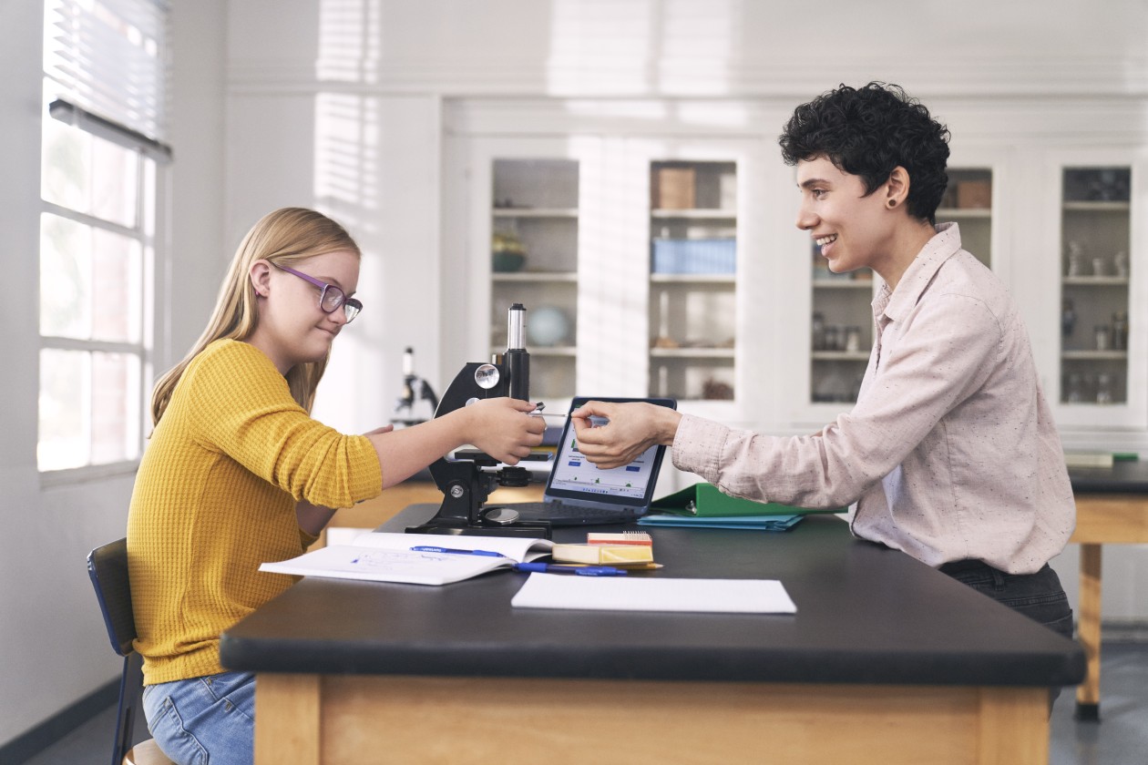 A student works with a microscope and a laptop in a science classroom while an educator hands a microscope slide to her.