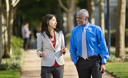 Image of two employees walking along a sidewalk discussing something over a mobile device.