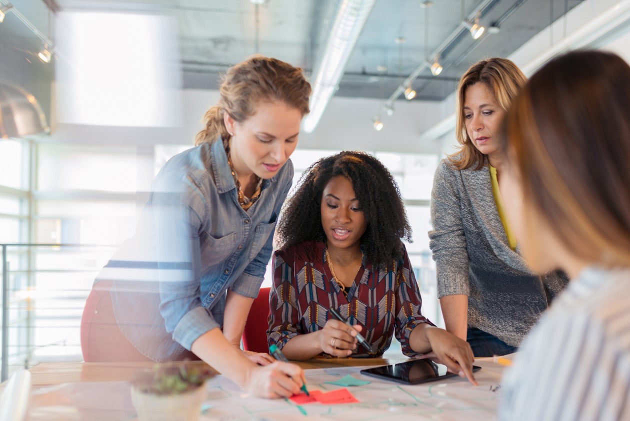 A diverse group of businesswomen look at a digital tablet and discuss a project in their modern office.