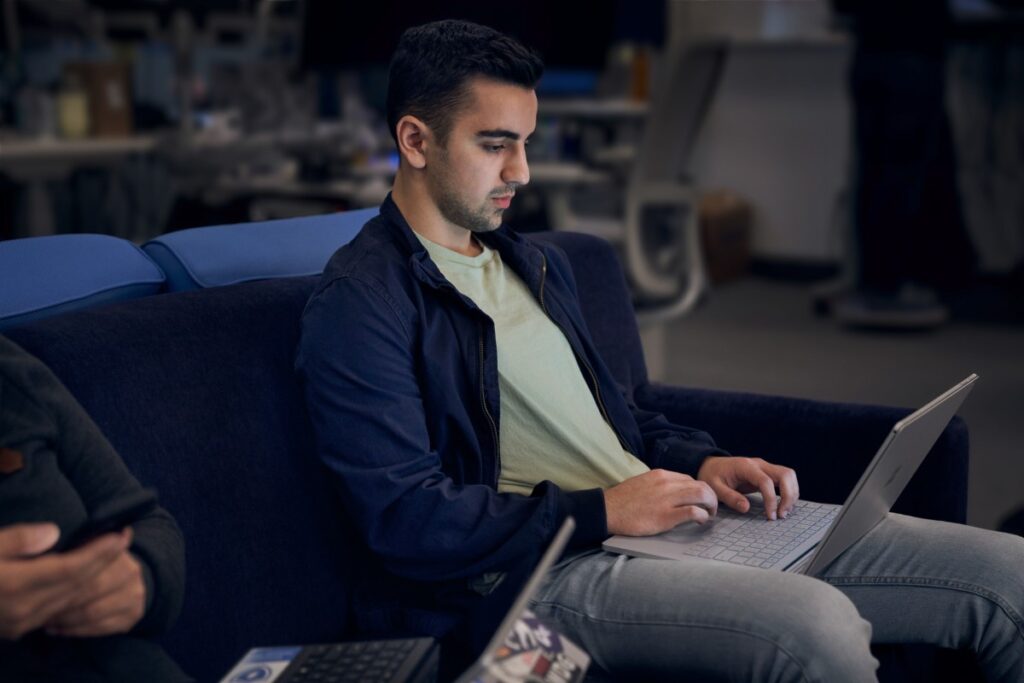 Lightly bearded man working on a laptop while sitting next to another person on a dark blue couch.