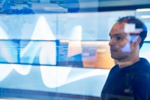 Man in sweater inside a secure room who is looking at data and a geographic area displayed on a large monitor which is behind glass walls with reflections.